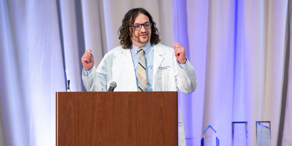 Graduate, Joey Salmone, wearing white lab coat and speaking at podium with awards on the table in the background