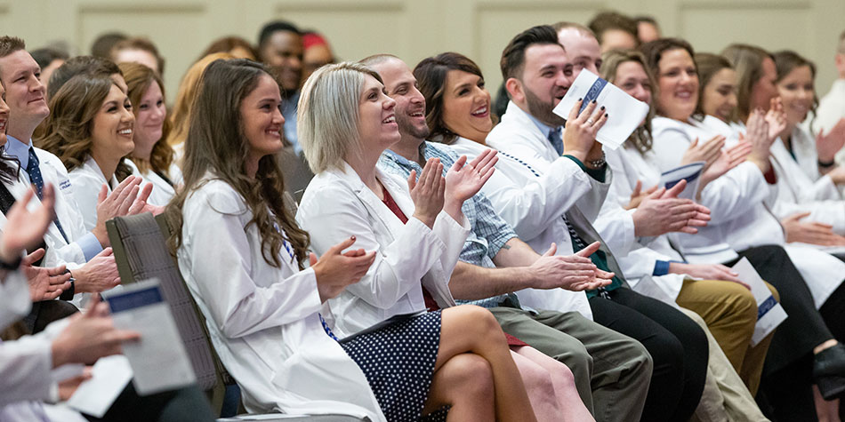 Seated graduates in the audience wearing white coats smiling and clapping while looking up at the stage