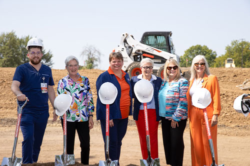 Large group of smiling people wearing hard hats and holding shovels over dirt