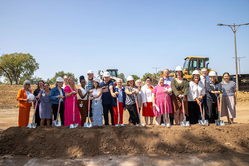 Small group of smiling people wearing hard hats and holding shovels over dirt