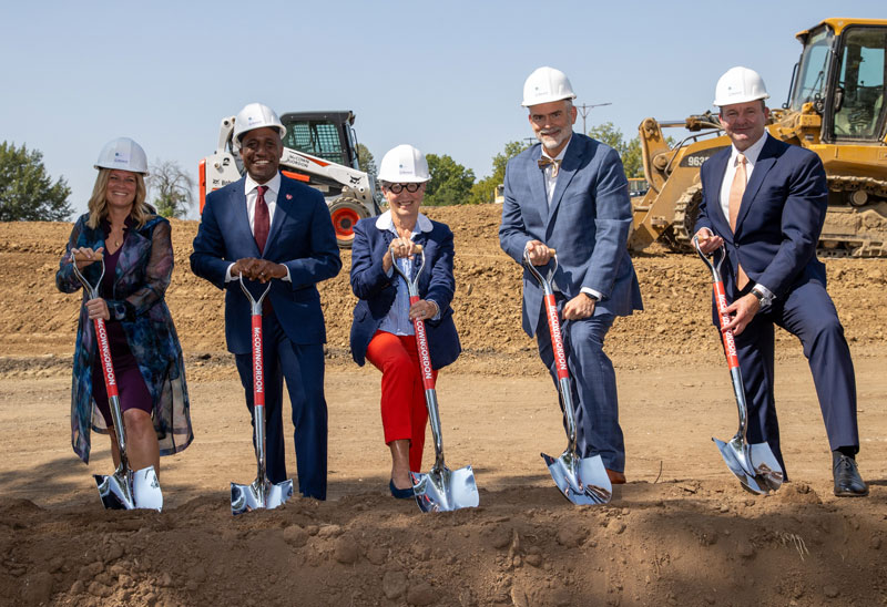 Small group of smiling people wearing hardhats and holding shovels over dirt