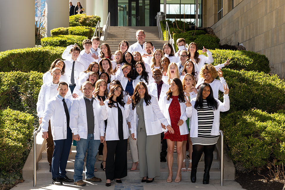 Group of graduates posing for a photo on the stairs outside a building wearing their white coats.