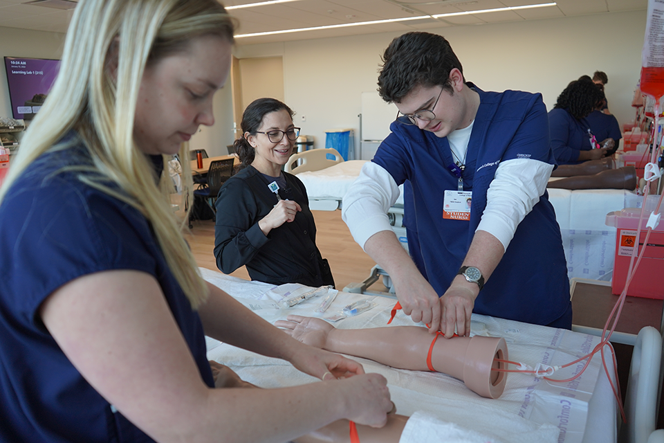 Professor guiding two students on how to draw blood on mannequin arms in the sim lab.