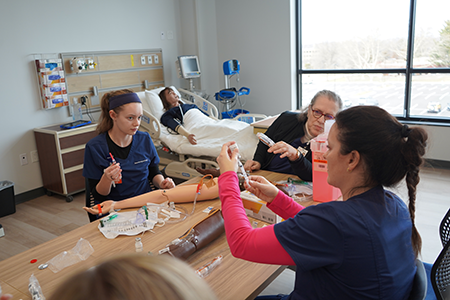 Students being taught to insert an IV by a professor in the sim suite.