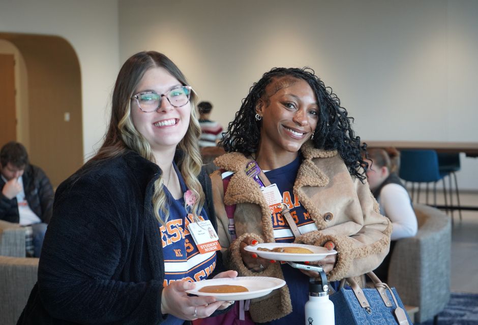 Two students in navy college‑themed shirts and jackets smile while holding a plate with a pancake, standing together in student lounge.