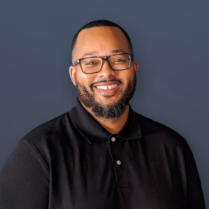 Portrait of Derick Jolliff-Cunigan wearing a black collared polo shirt and glasses.