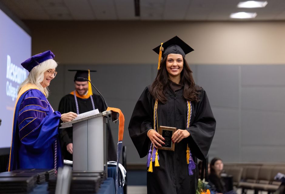 Student Riley Fate wearing graduation cap and gown walking across stage with award.